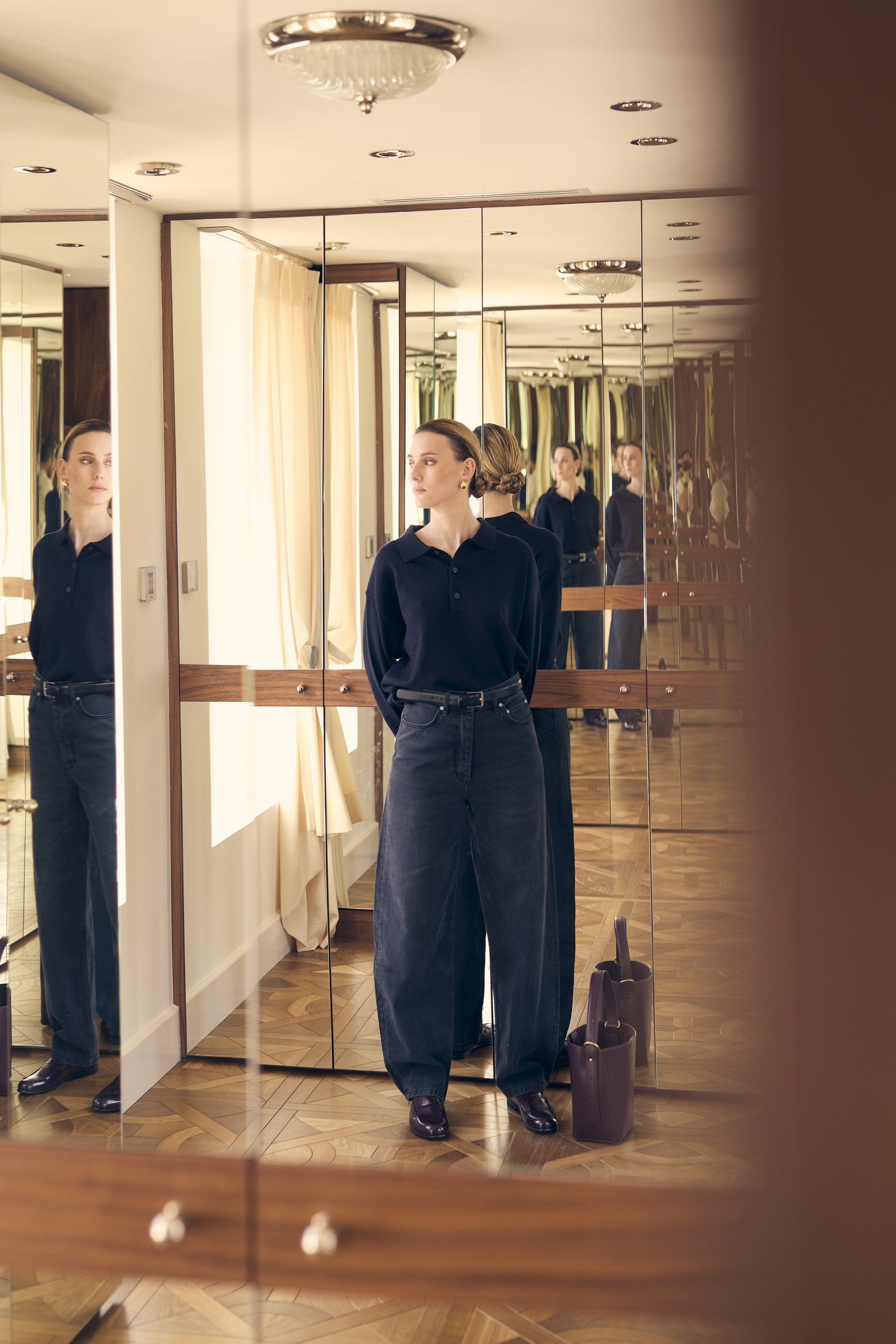 Woman standing in a room with reflective surfaces and a suitcase.