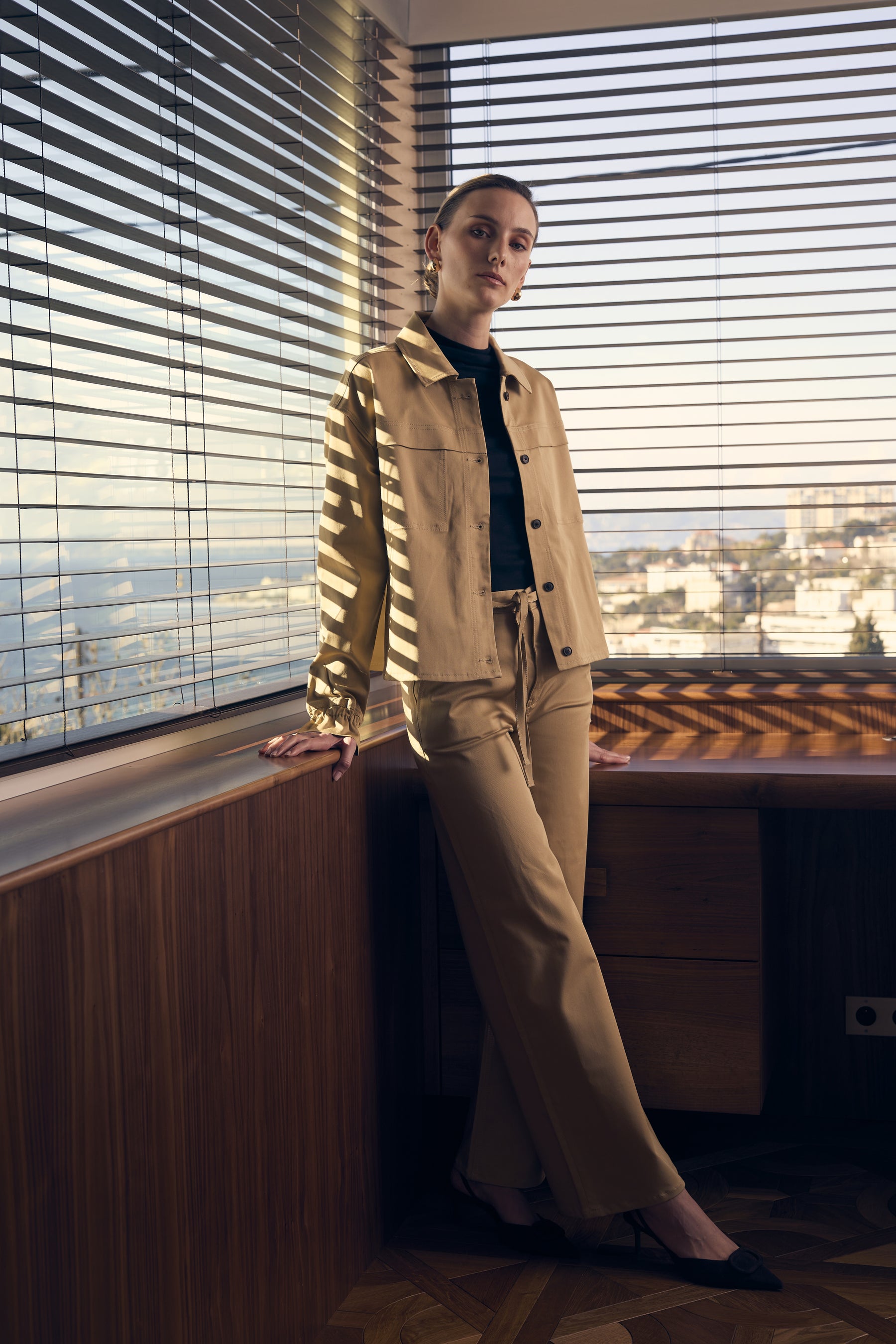 Woman in beige jacket and pants standing by a window with blinds.