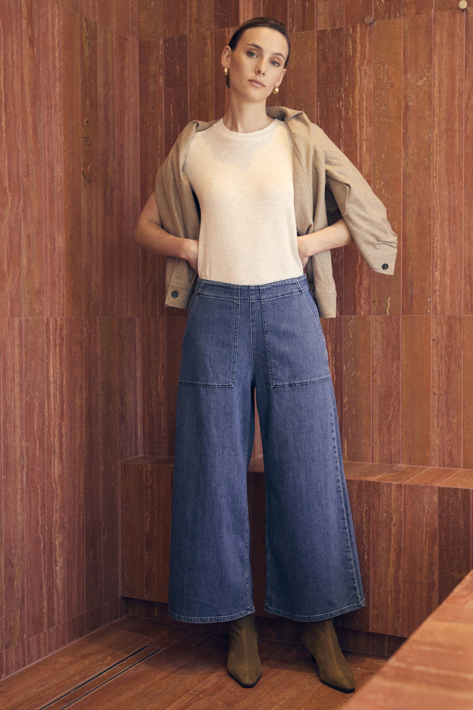 Person wearing a beige jacket, white shirt, and blue jeans standing against a wooden paneled wall.
