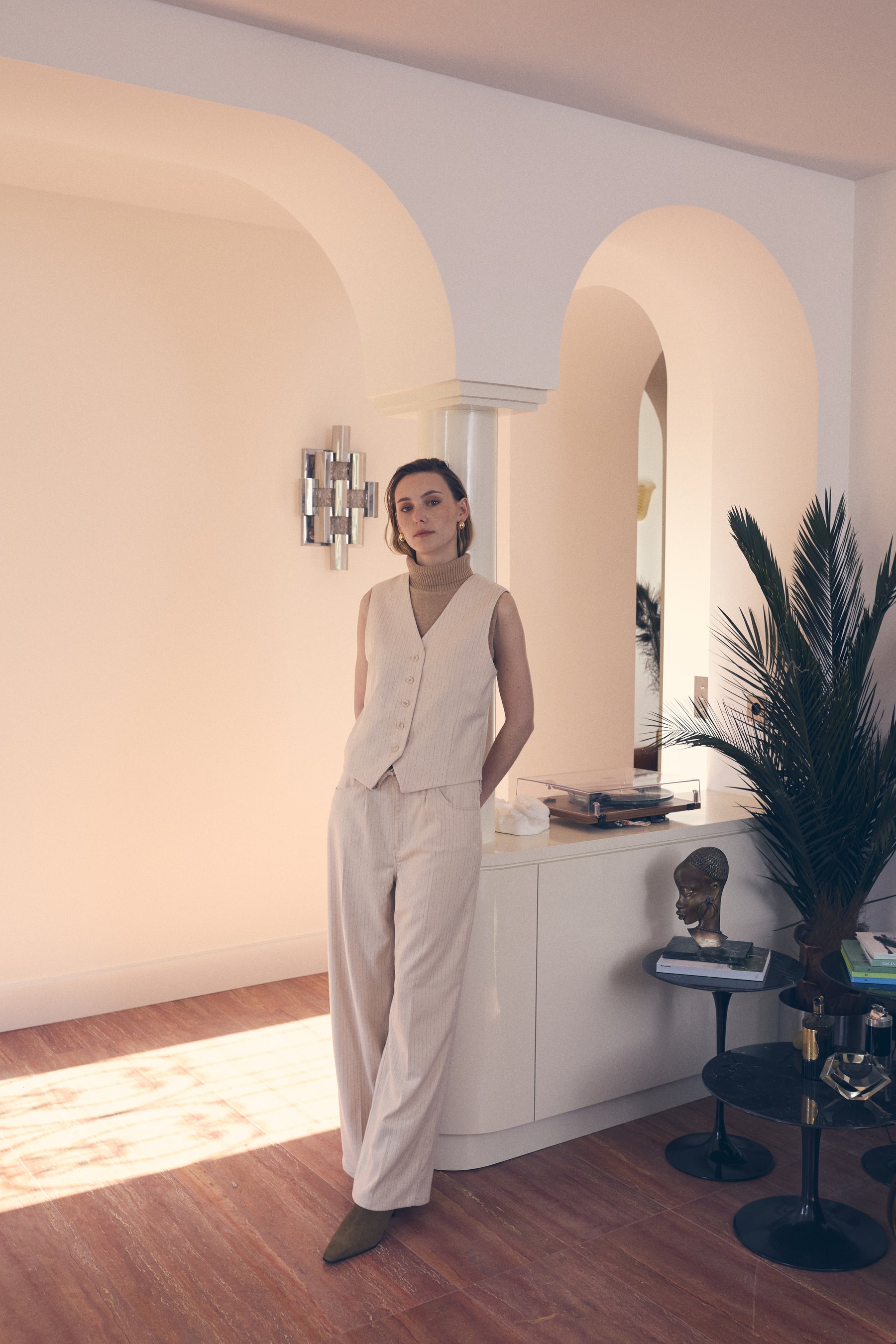 Woman in a beige outfit standing in a modern interior setting with arches and decor.