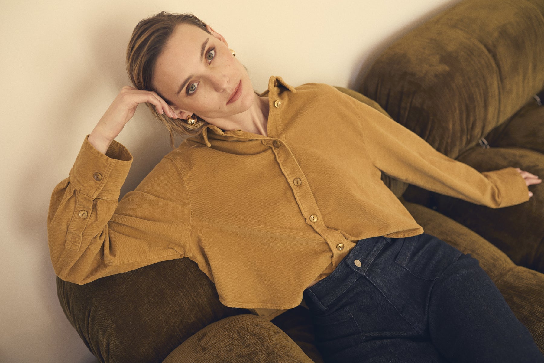 Woman wearing a mustard yellow shirt and blue jeans sitting on a brown couch.