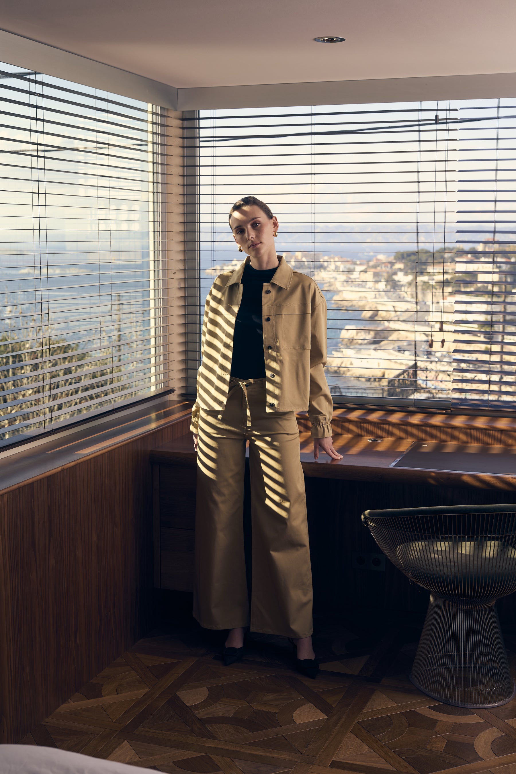 Person in a beige outfit standing in a room with large windows and a cityscape view.