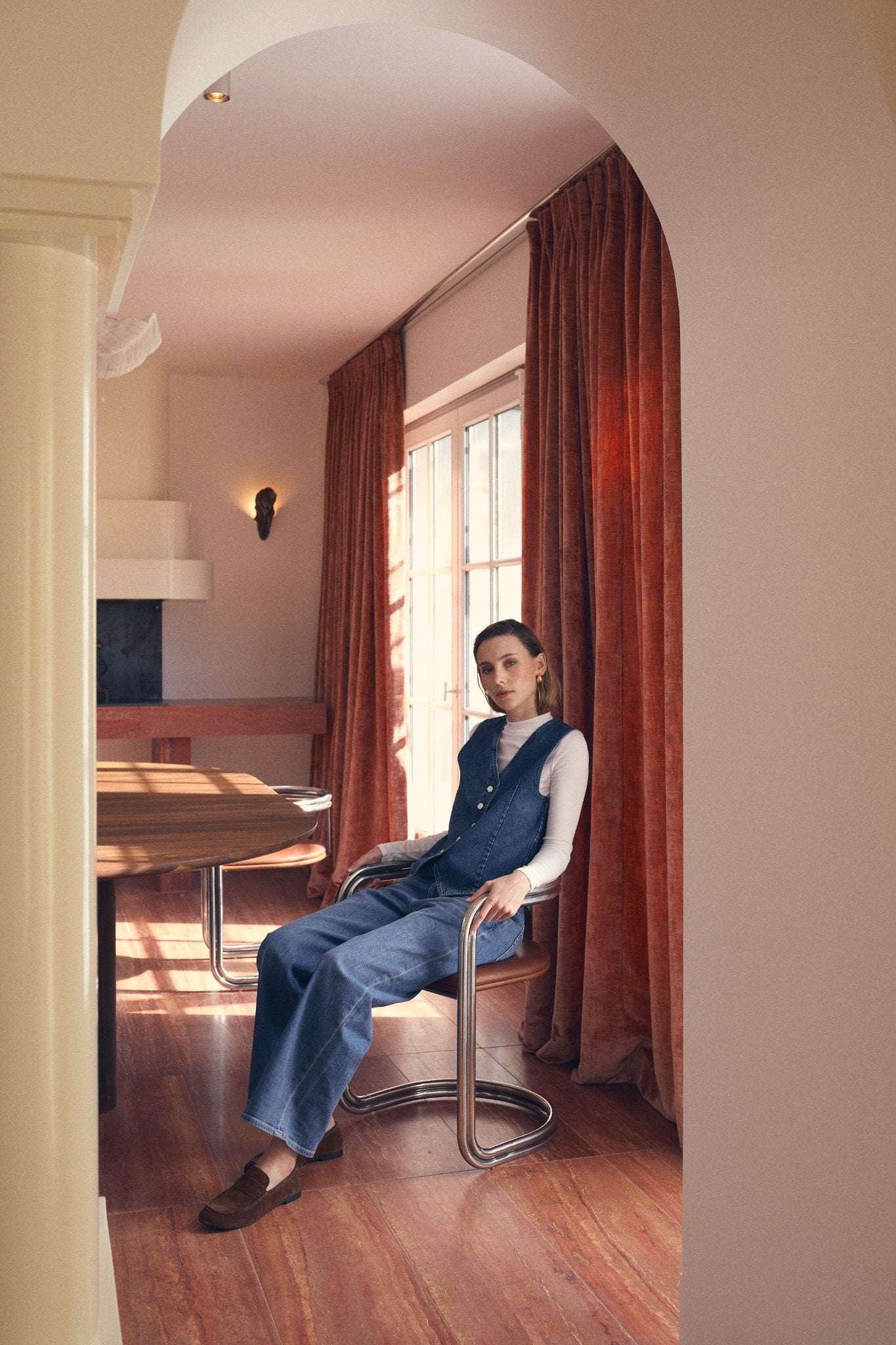 Woman sitting in a modern dining room wearing a denim vest, wide-leg jeans, white long-sleeve shirt, and brown loafers.